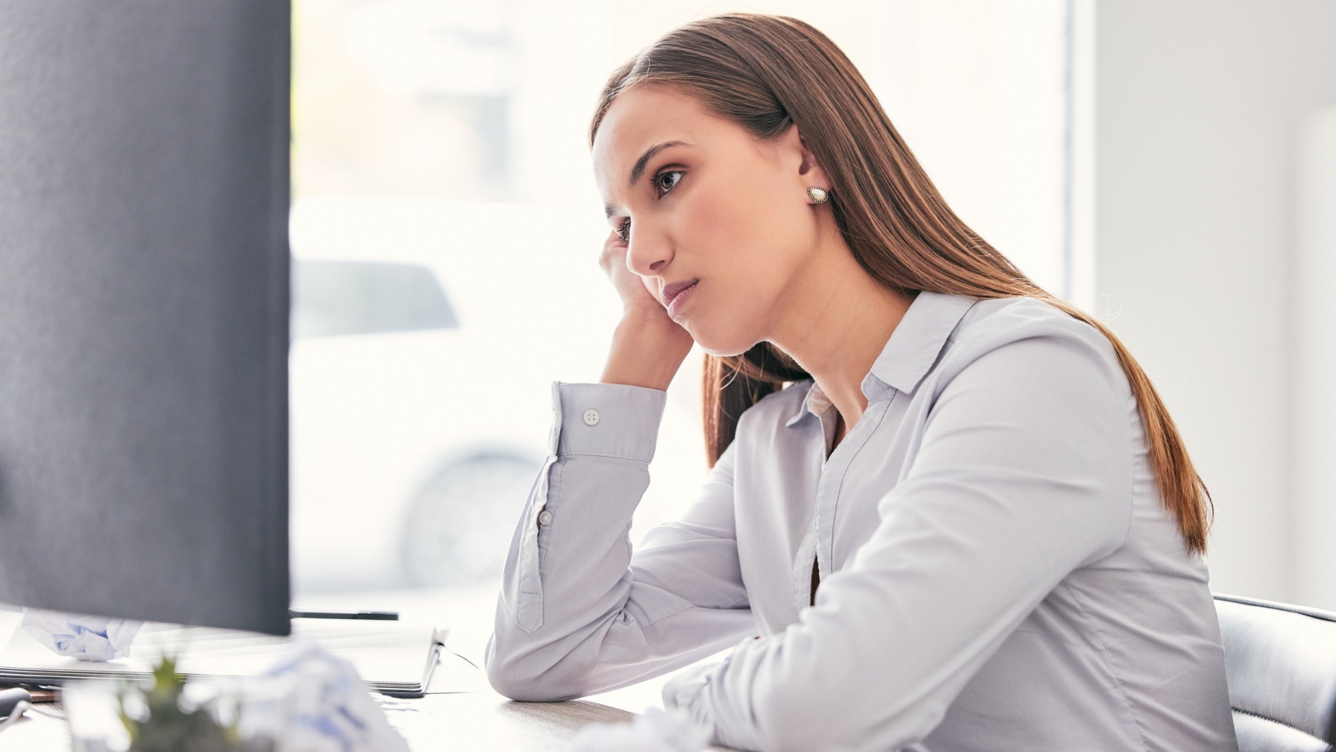 A woman sits at her desk, propping her head on her hand, looking unenthusiastic as she stares at her computer.